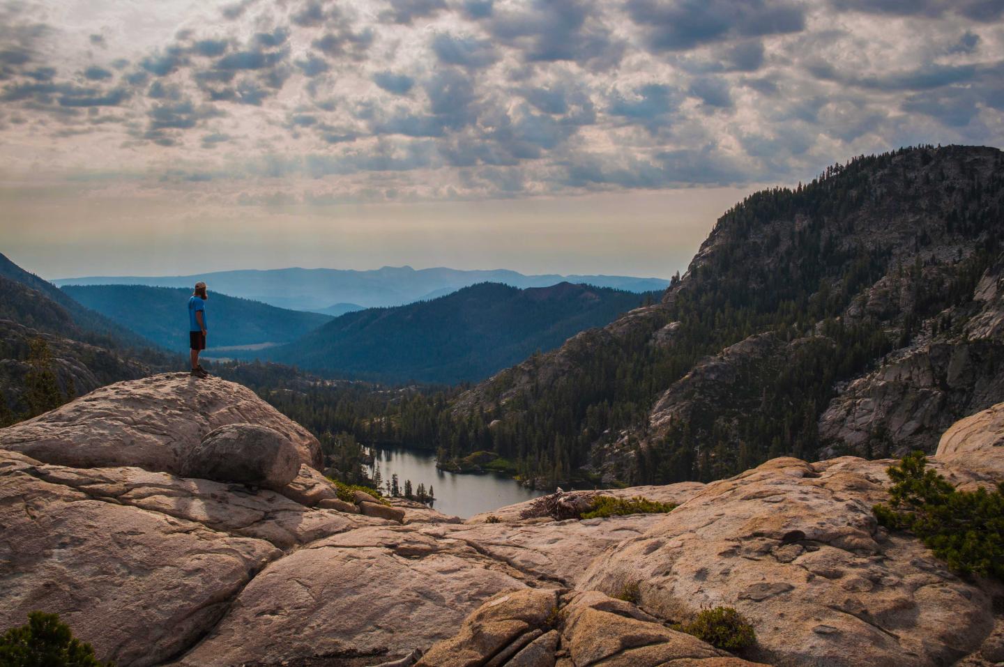 Man stands on rocky outcrop overlooking scenic mountain view with cloudy sky.