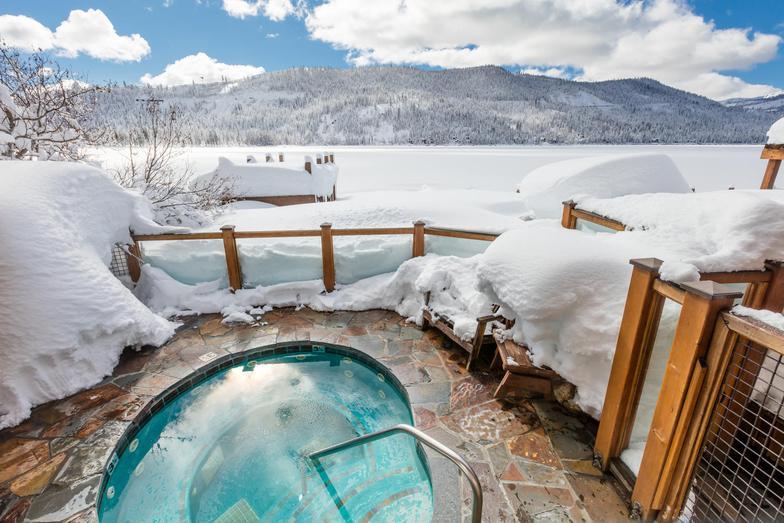 Snow-covered hot tub overlooking a frozen lake and snowy mountains.