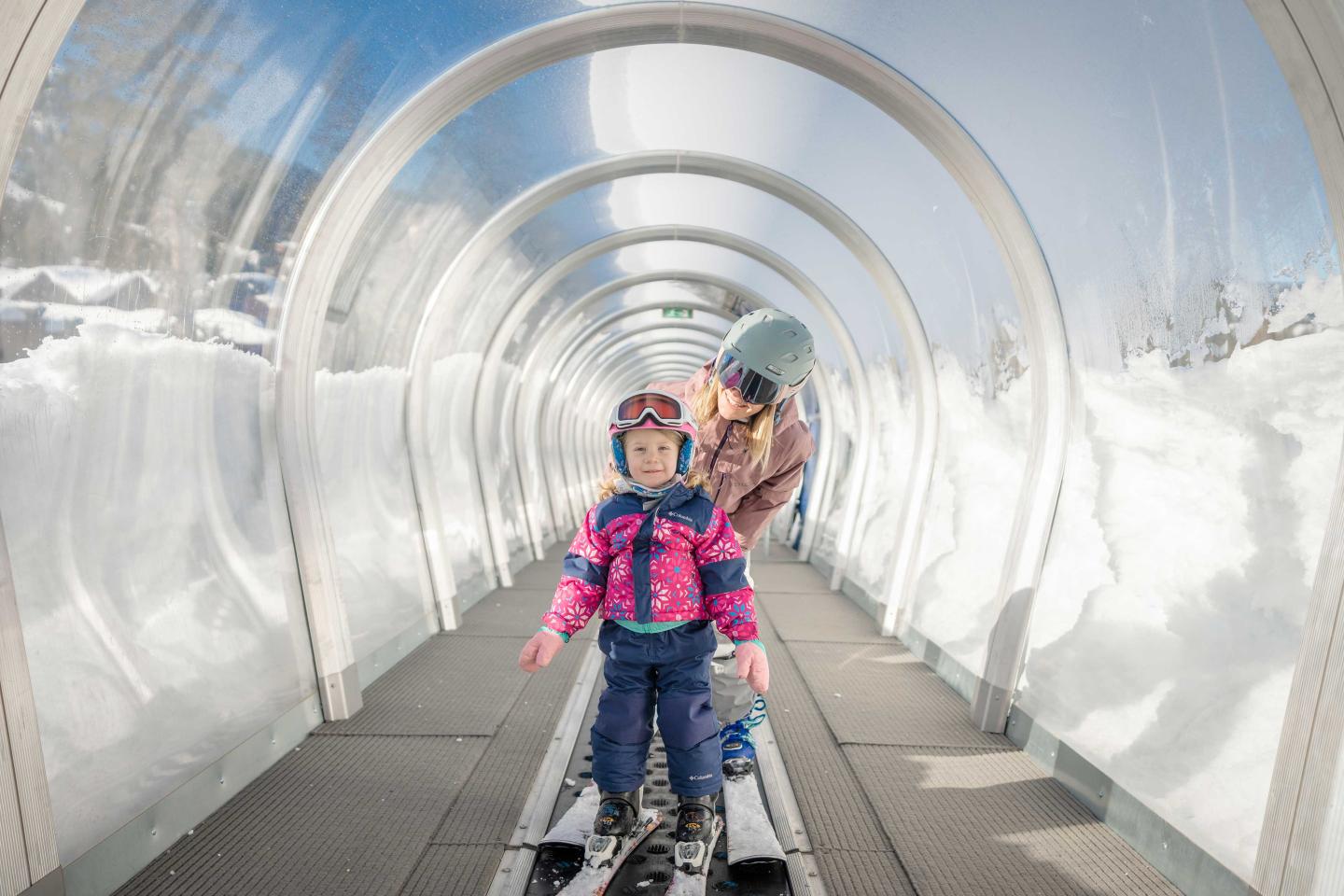 Child skiing on conveyor belt in snow tunnel, guided by an adult.