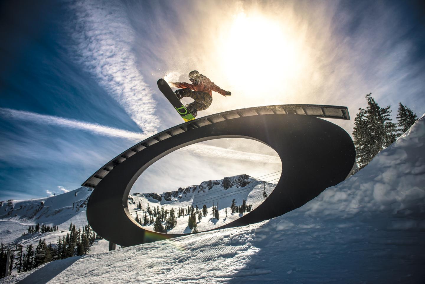 Snowboarder jumping over a circular ramp against a bright sunlit sky.