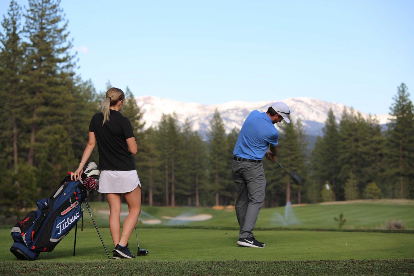 Golfer swings on green, woman watches, mountains in background.
