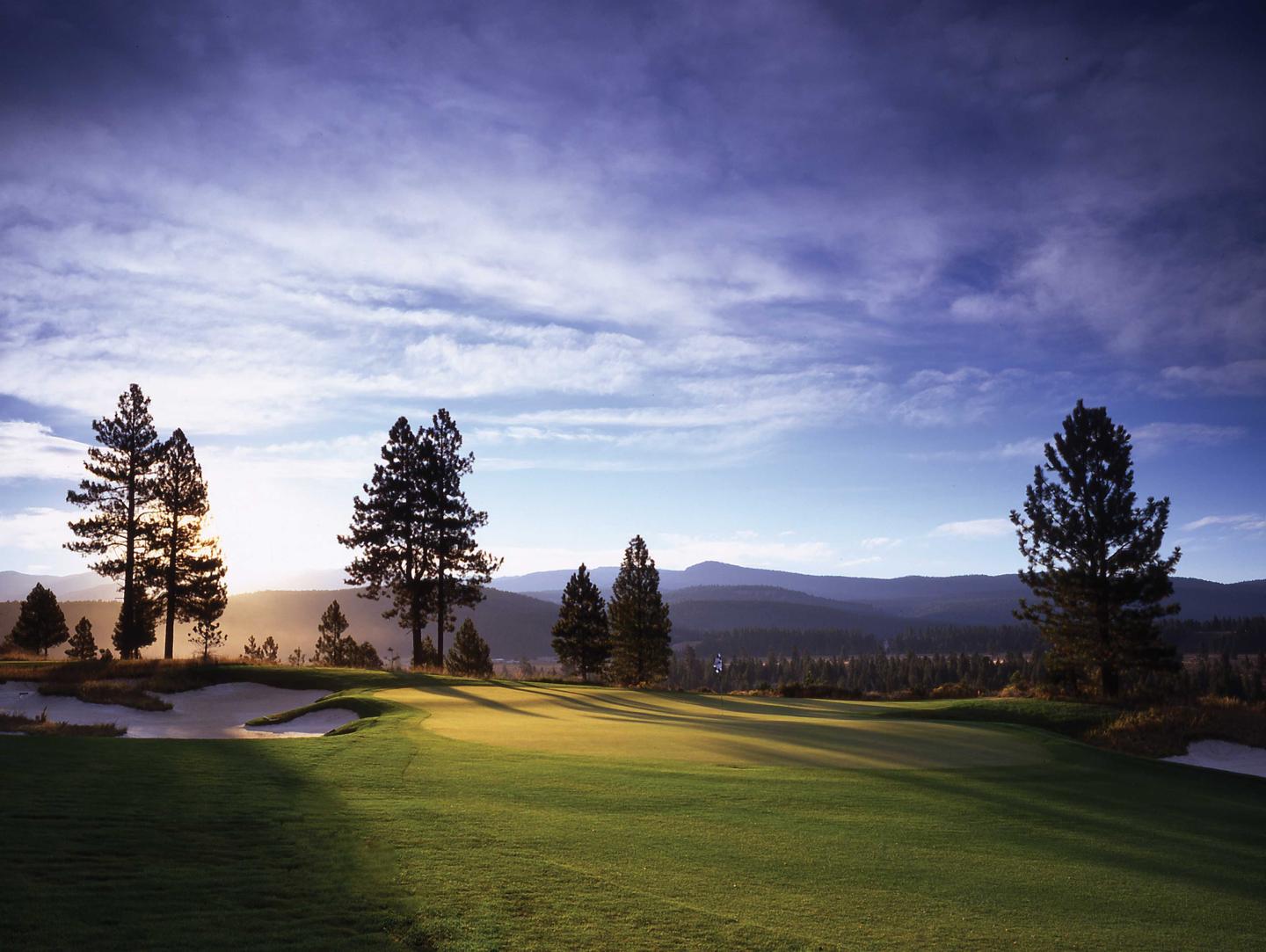 Golf course at sunrise with trees and mountains in the background.