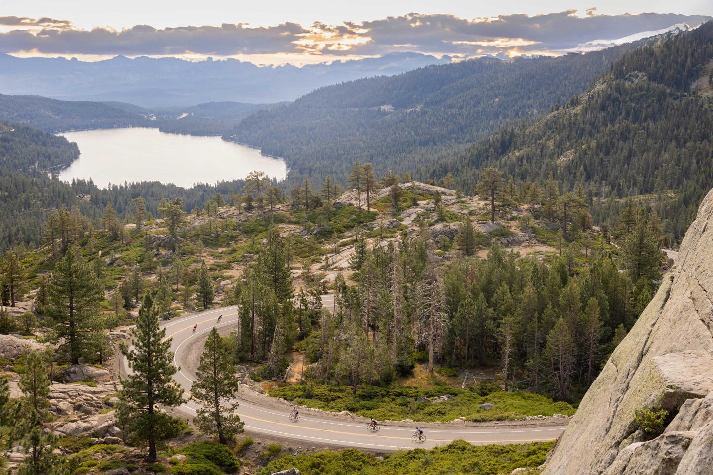 Mountain landscape with winding road, pine trees, and lake under a cloudy sky.