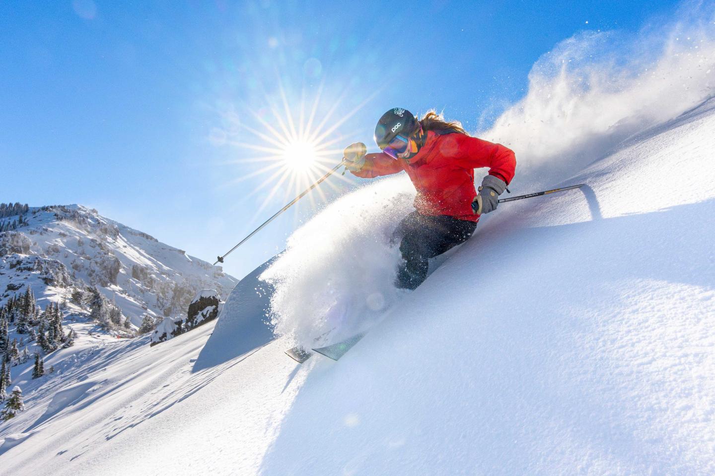 Skier in red jacket descends snowy slope under bright sun.