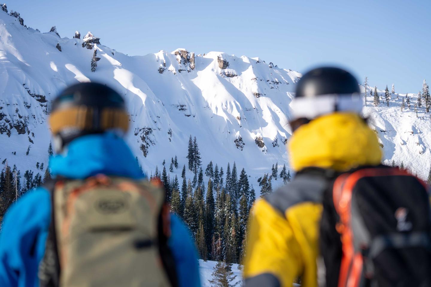 Two skiers in helmets facing snowy mountains in bright sunlight.