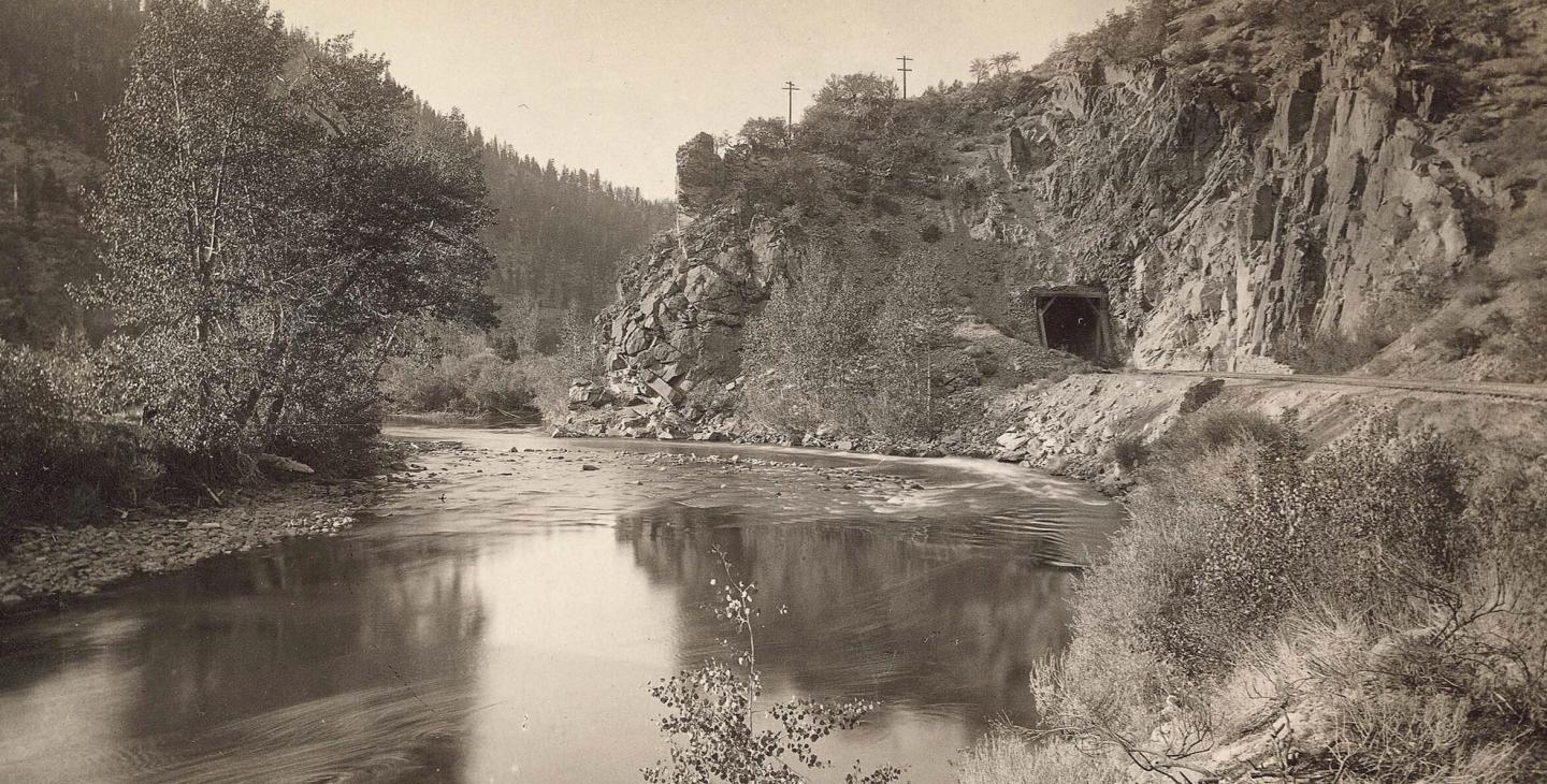 River winding through rocky canyon with trees and small tunnel entrance. Sepia photograph.