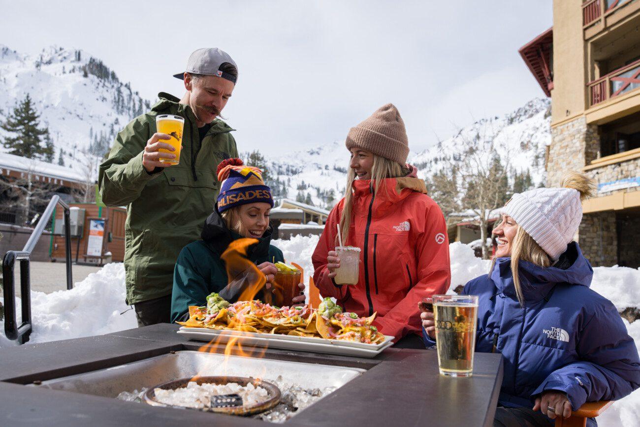 Friends enjoying drinks and nachos by a fire pit in a snowy mountain setting.