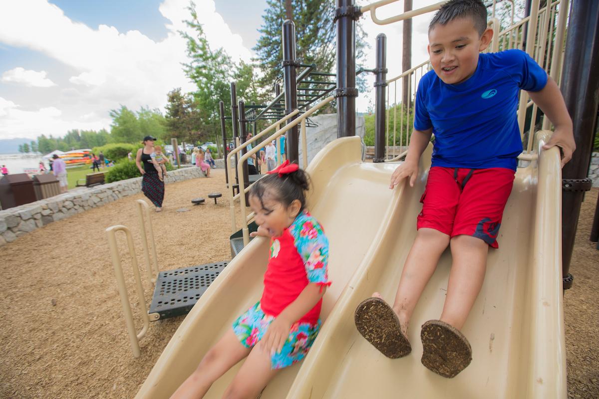 Children smiling while sliding down a playground slide in summer clothing.