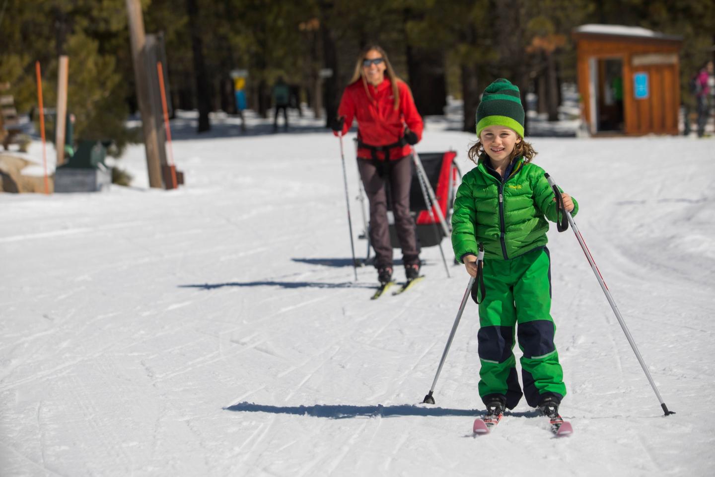 Tahoe Donner Child in green skiing, followed by adult in red, on a snowy trail amid trees.