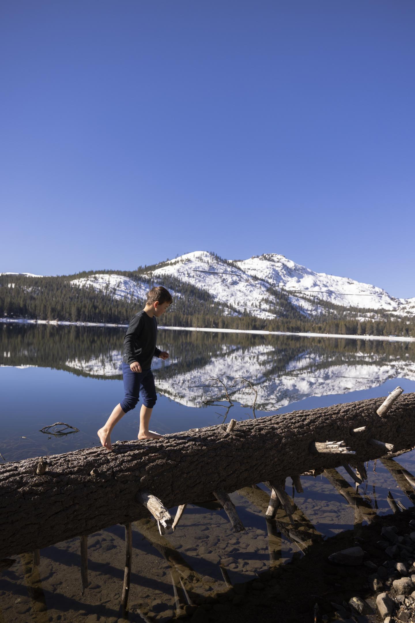 Barefoot kid walks on a log over Donner Lake, snowy mountain in the background in the spring