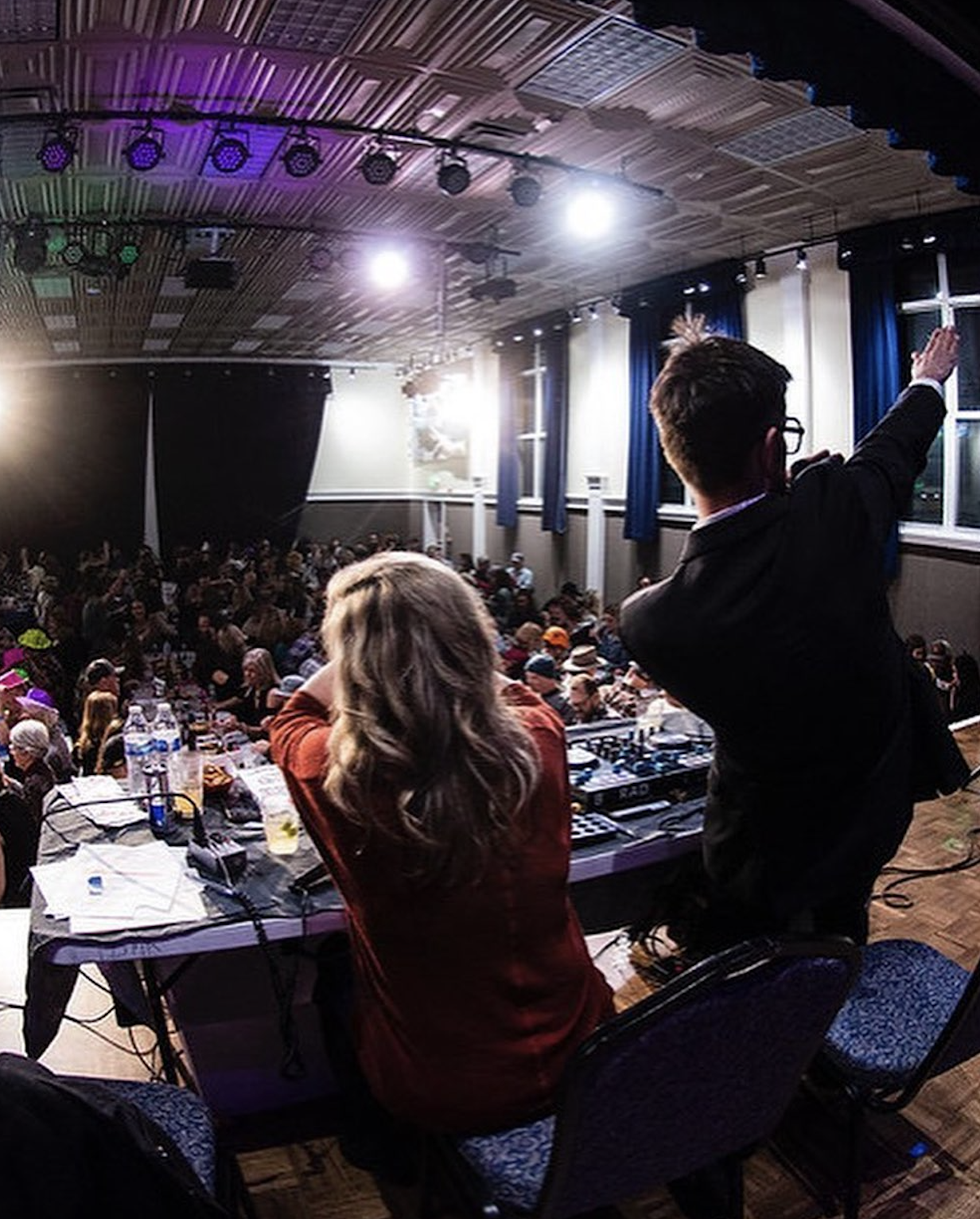 Two presenters on stage facing an audience in a brightly lit auditorium.