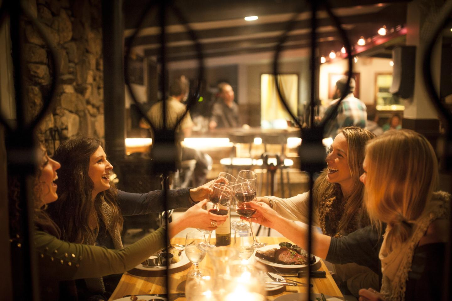 Four women clink wine glasses at a cozy restaurant table, smiling and enjoying themselves.