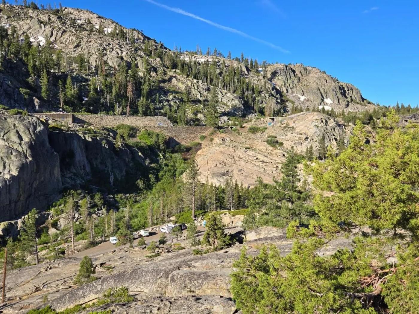 Mountain landscape with rocky cliffs, green trees, and a clear blue sky.