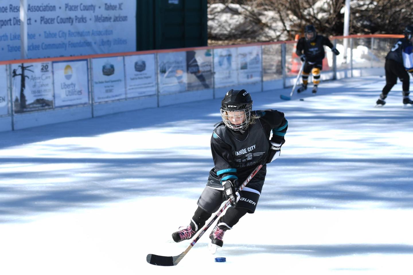Child playing hockey on an outdoor rink, wearing black gear and skating with a stick.