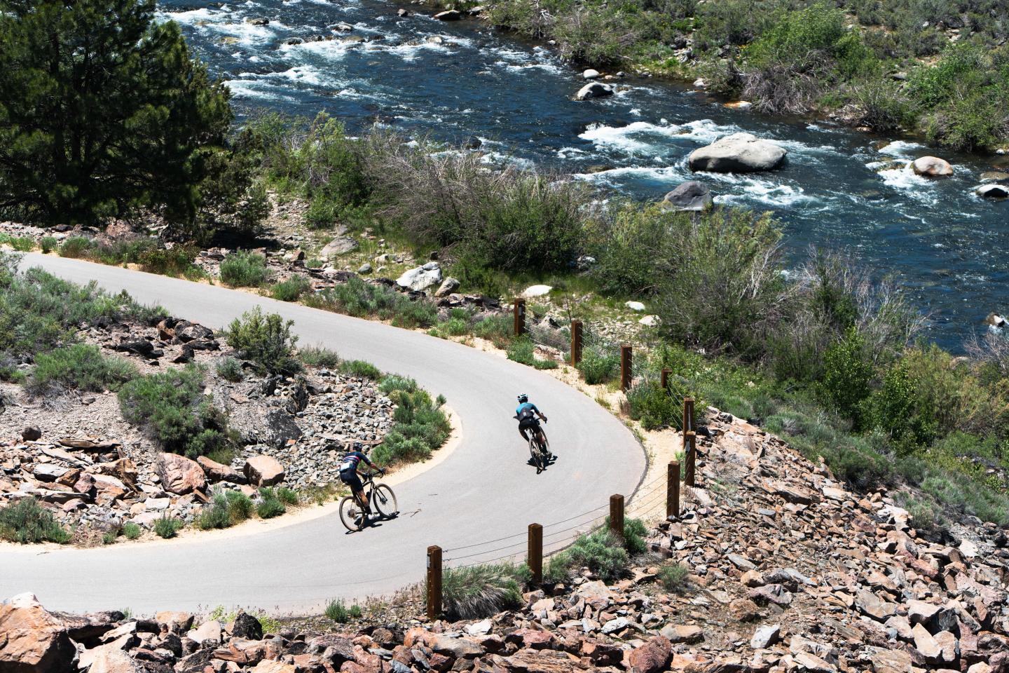 Cyclists on a winding road by a flowing river.