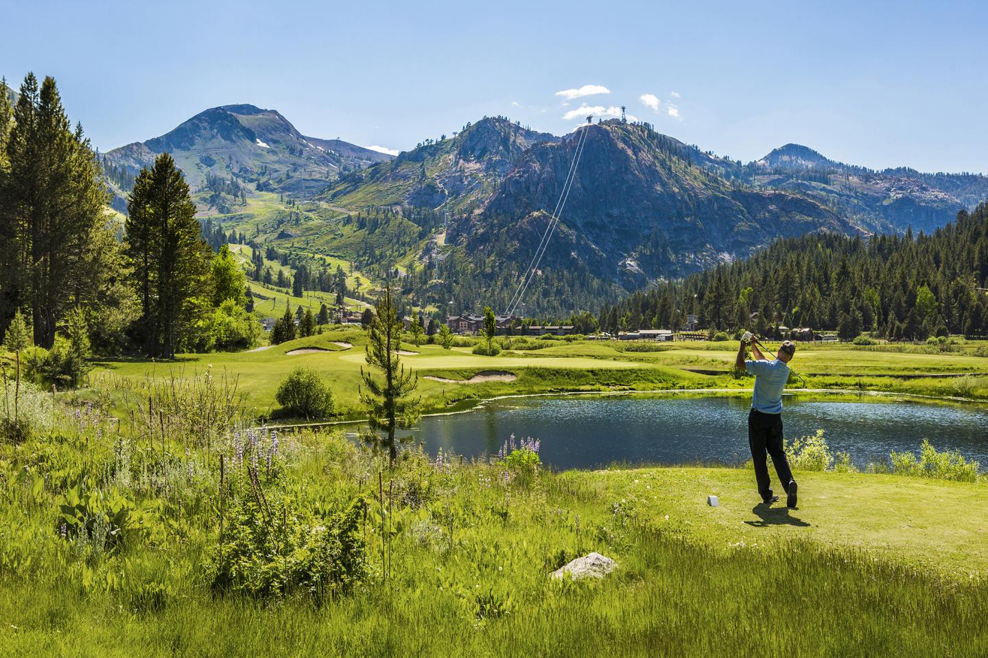 Golfer swings on a lush, mountain-backed course under a clear blue sky.
