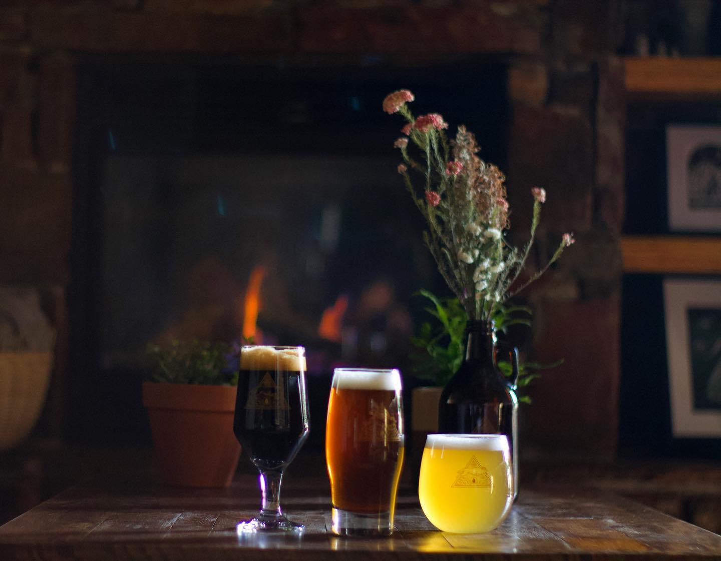 Three glasses of beer and flowers on a wooden table, cozy fireplace background.