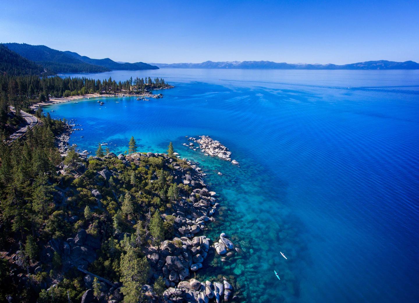 Aerial view of a turquoise lake surrounded by pine forests and mountains under a clear blue sky.