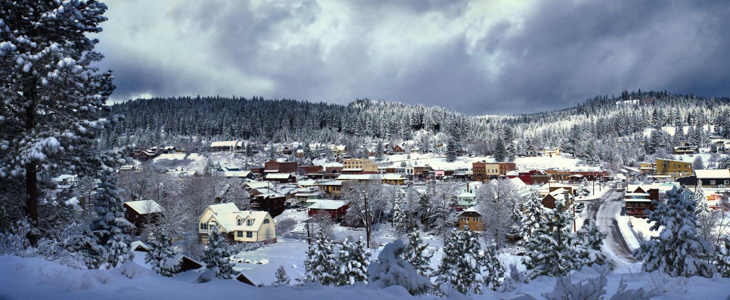 Snowy town under cloudy sky, surrounded by snow-covered trees and hills.
