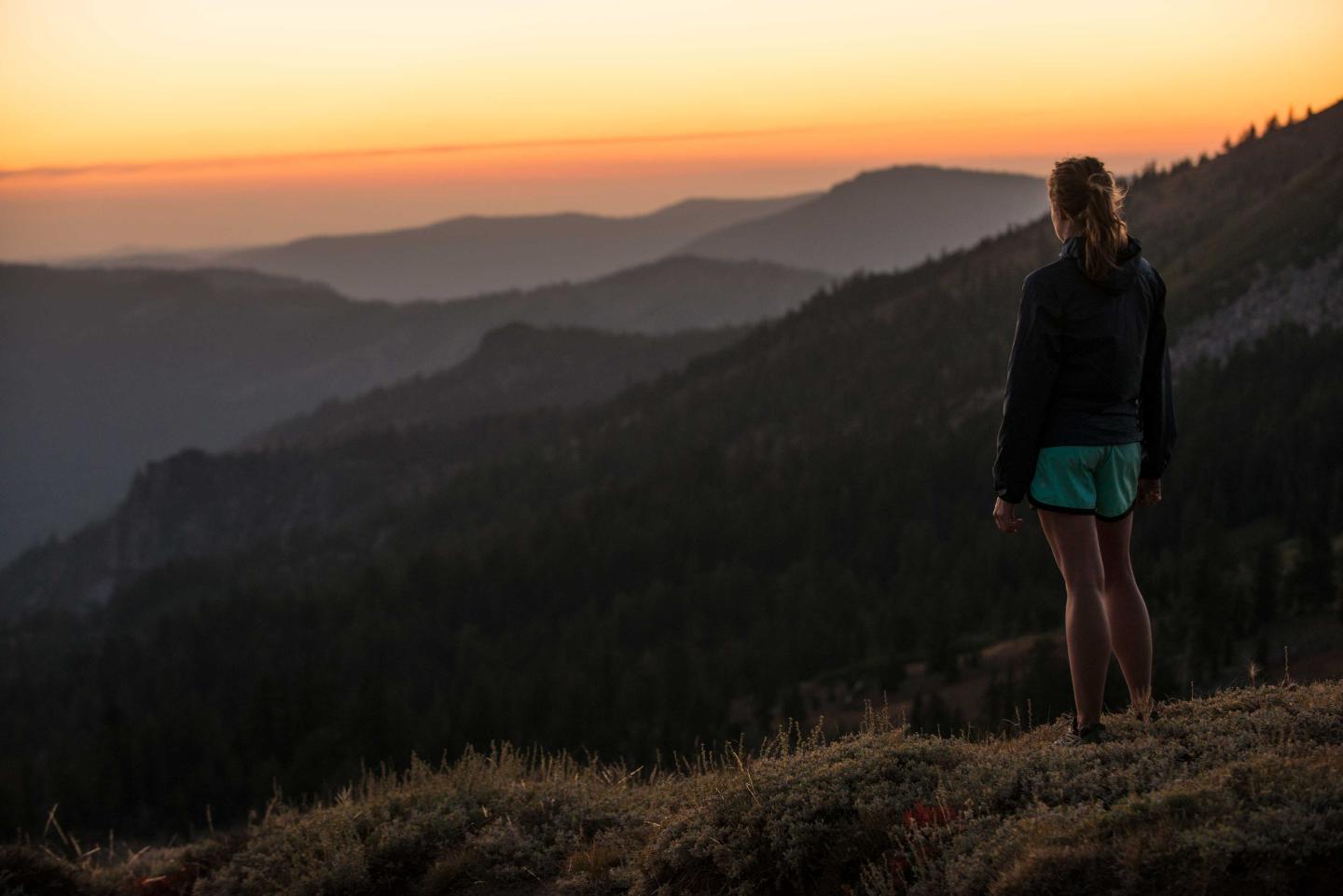 Woman standing on mountain, gazing at sunset over layered hills.