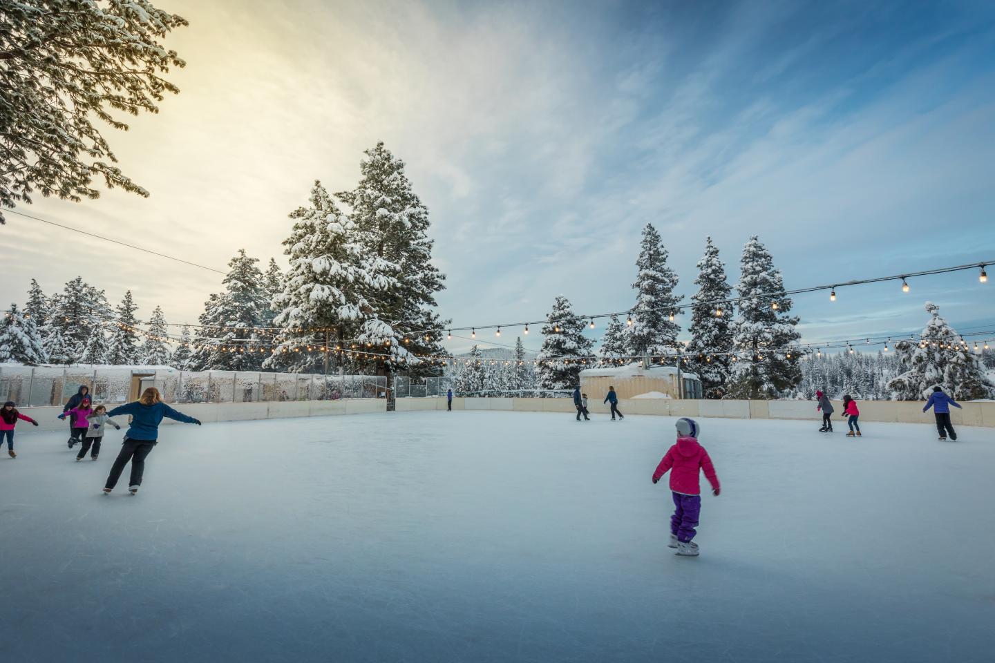 Outdoor ice rink with people skating, surrounded by snowy trees at sunset, Truckee, California