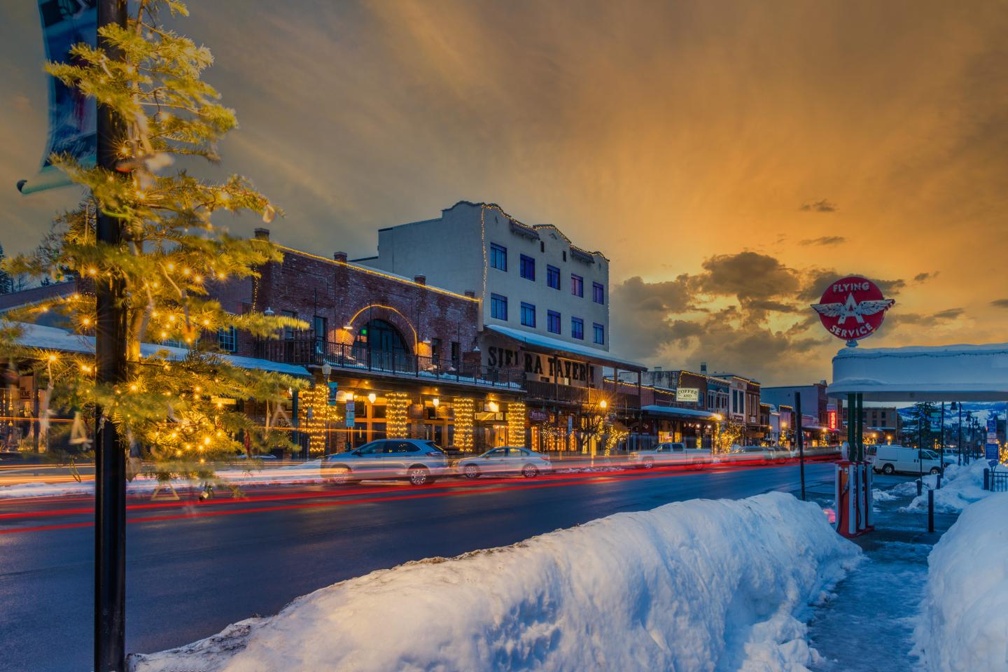 Snowy street at sunset with colorful lights and blurred car trails.