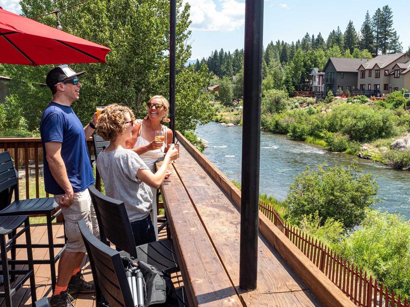People on a deck by a river, toasting with drinks, under a red umbrella.