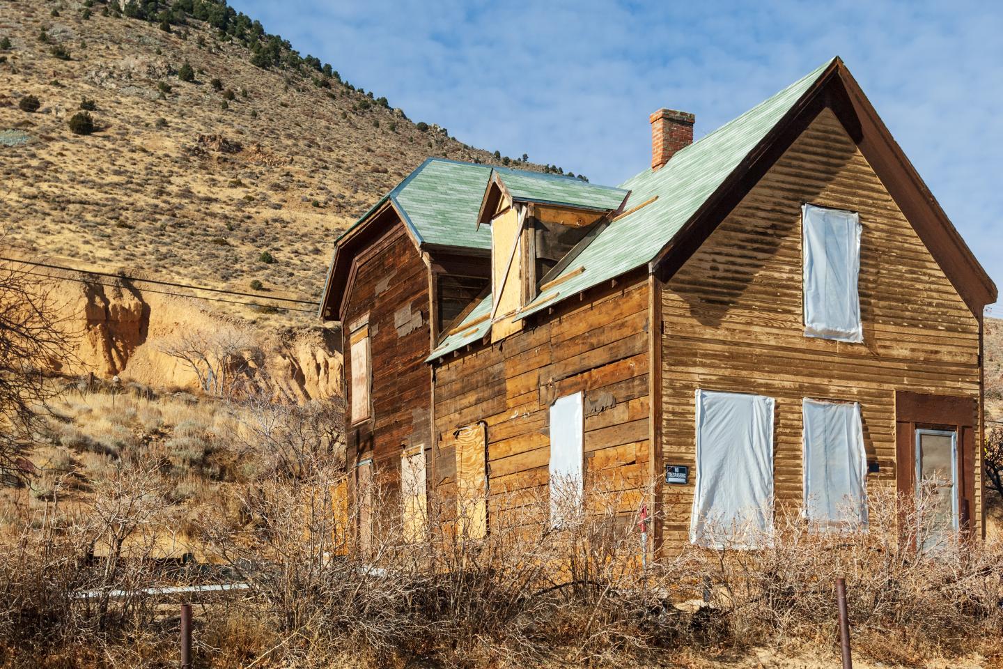 Old wooden house with boarded windows in a dry, mountainous landscape.