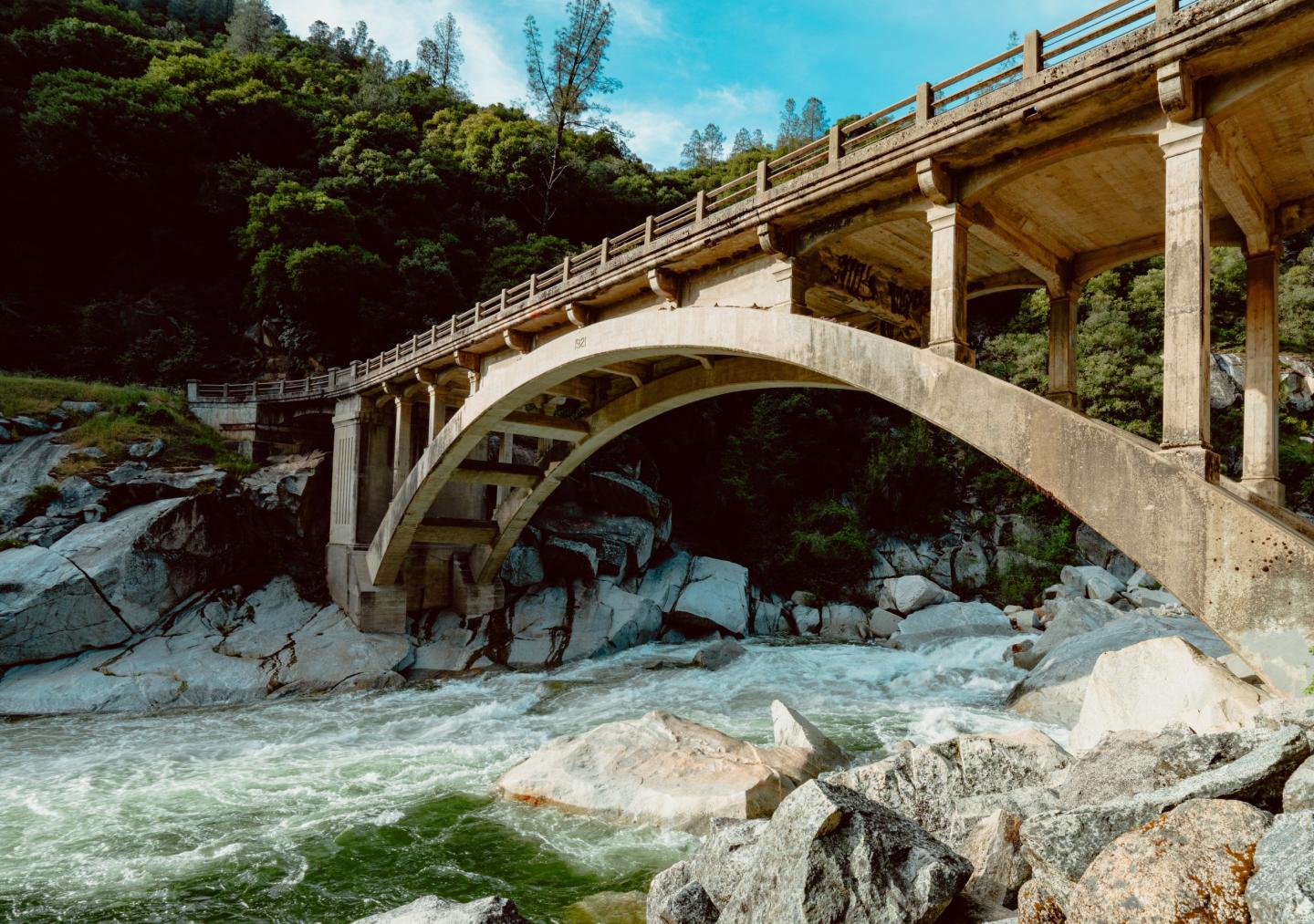 Concrete arch bridge over a flowing river, surrounded by greenery.
