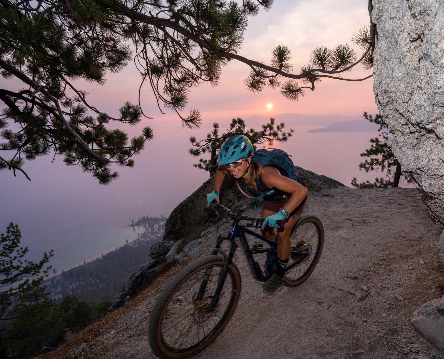 Mountain biker on rocky trail at sunset surrounded by trees and pink sky.