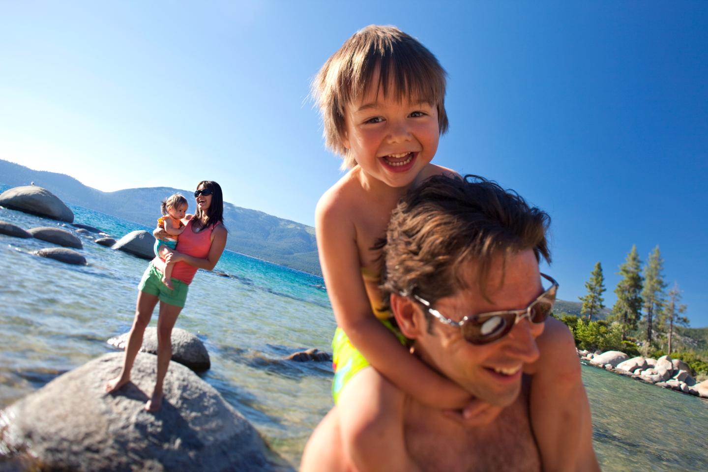 Family enjoying a sunny day by the lake; child on a man's shoulders, another with a woman.