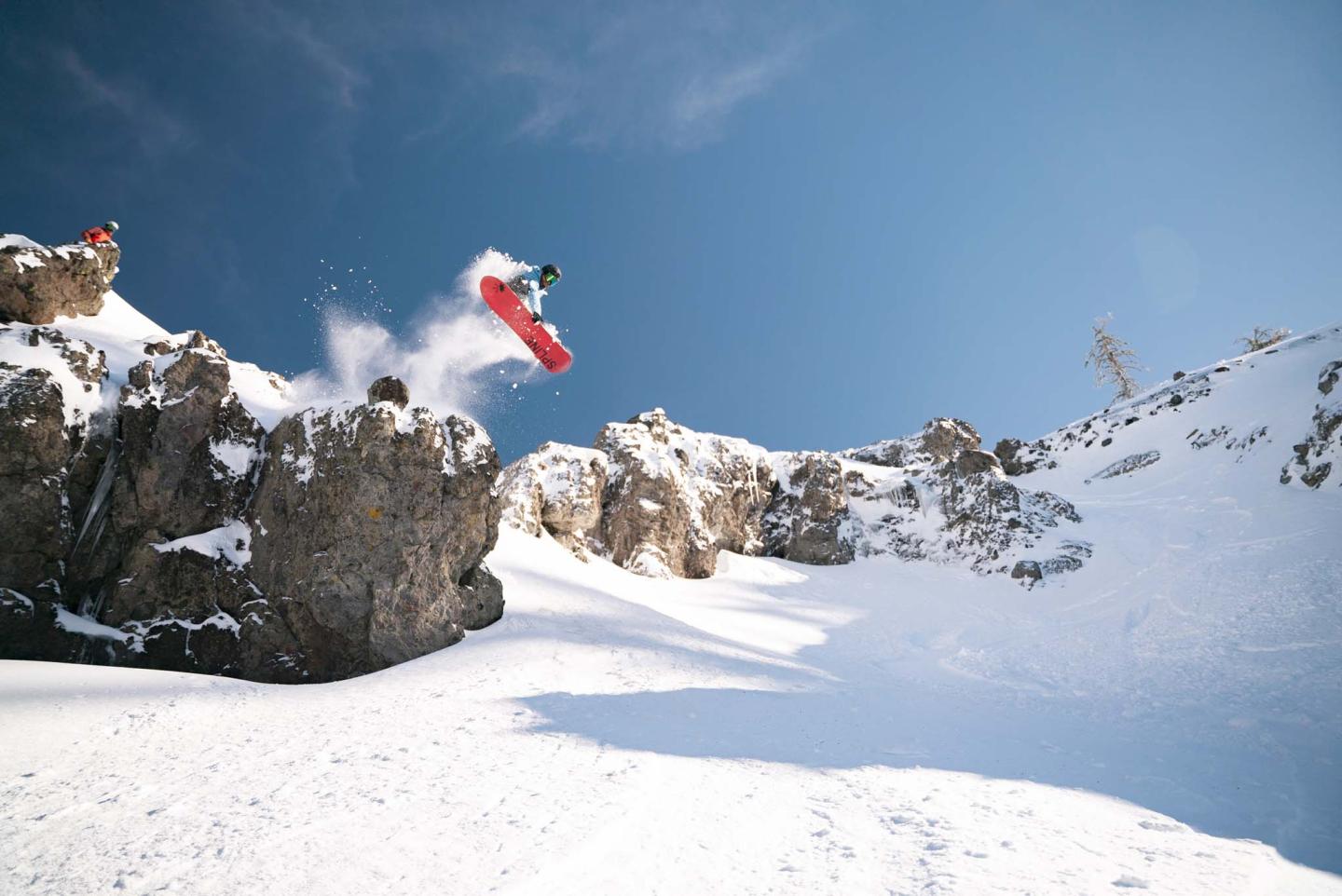Snowboarder jumping off a snowy cliff under a clear blue sky.