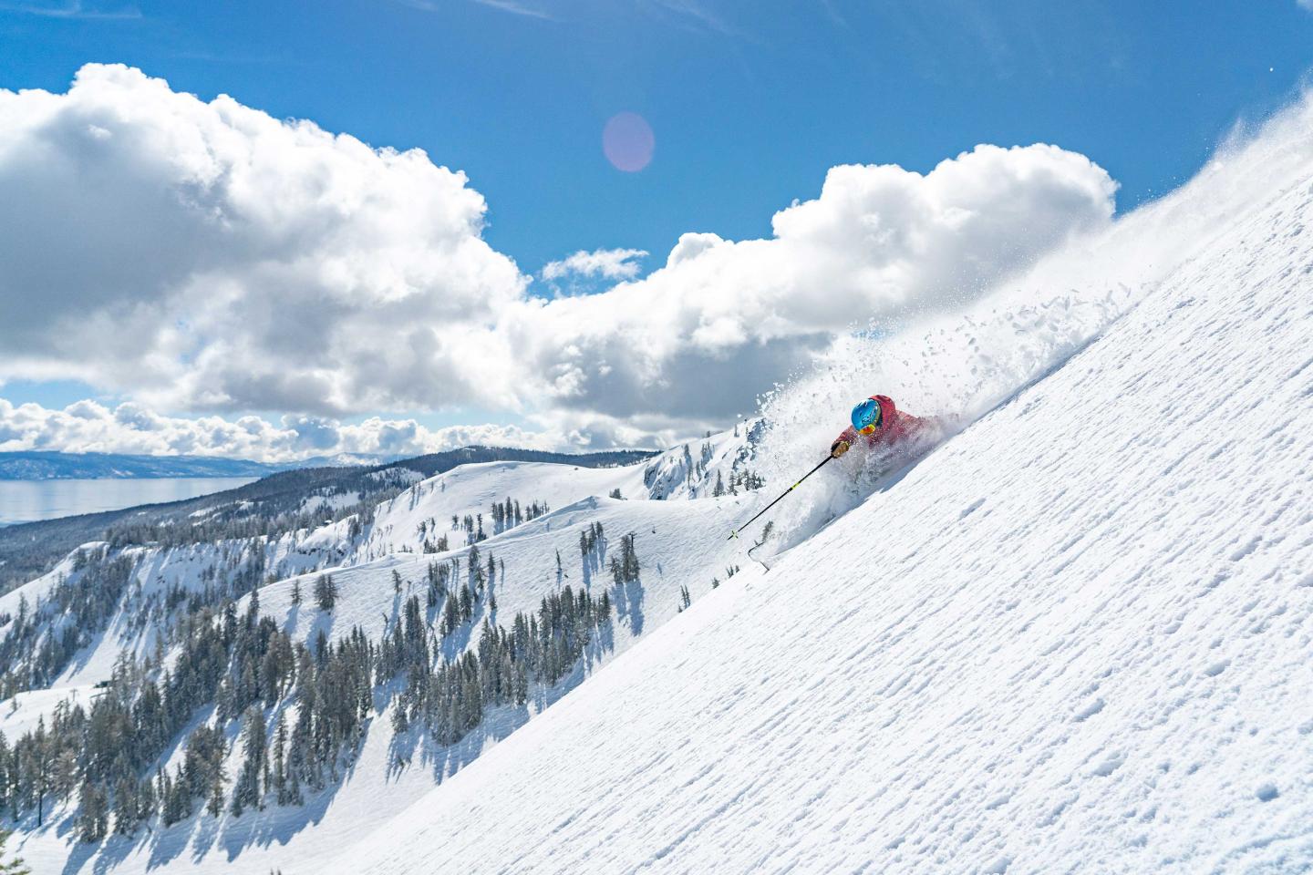 Skier in red descends snowy mountain under blue sky with clouds.