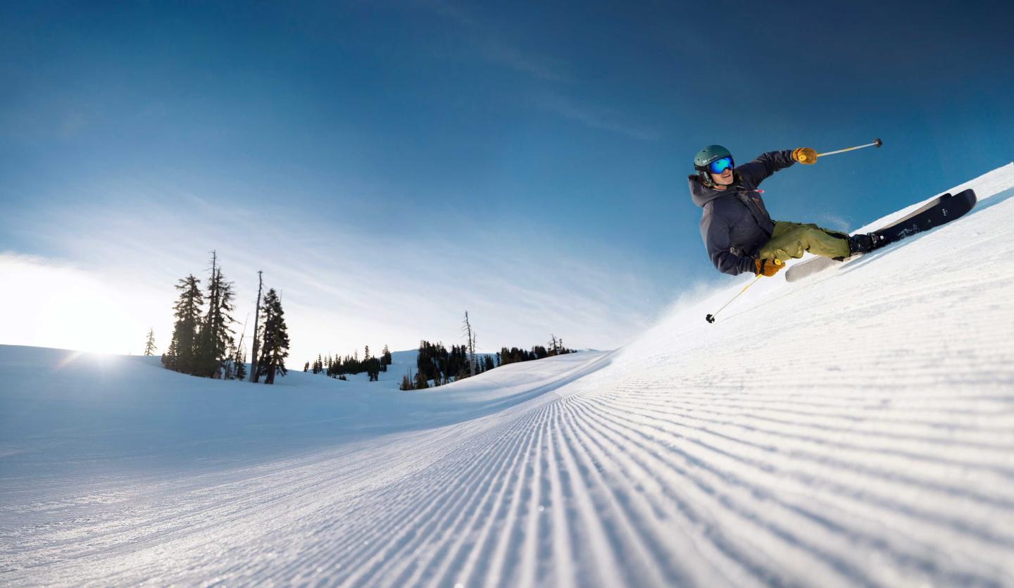 Skier carving down a sunlit snowy slope under a clear blue sky.