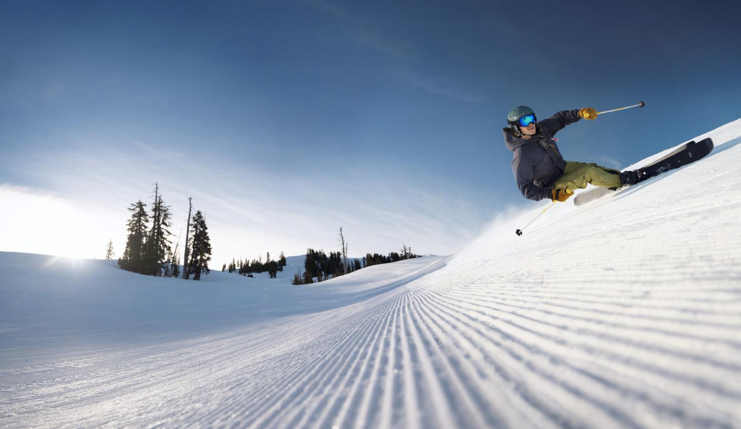 Skier carving through snow on a sunny slope with trees in the background.