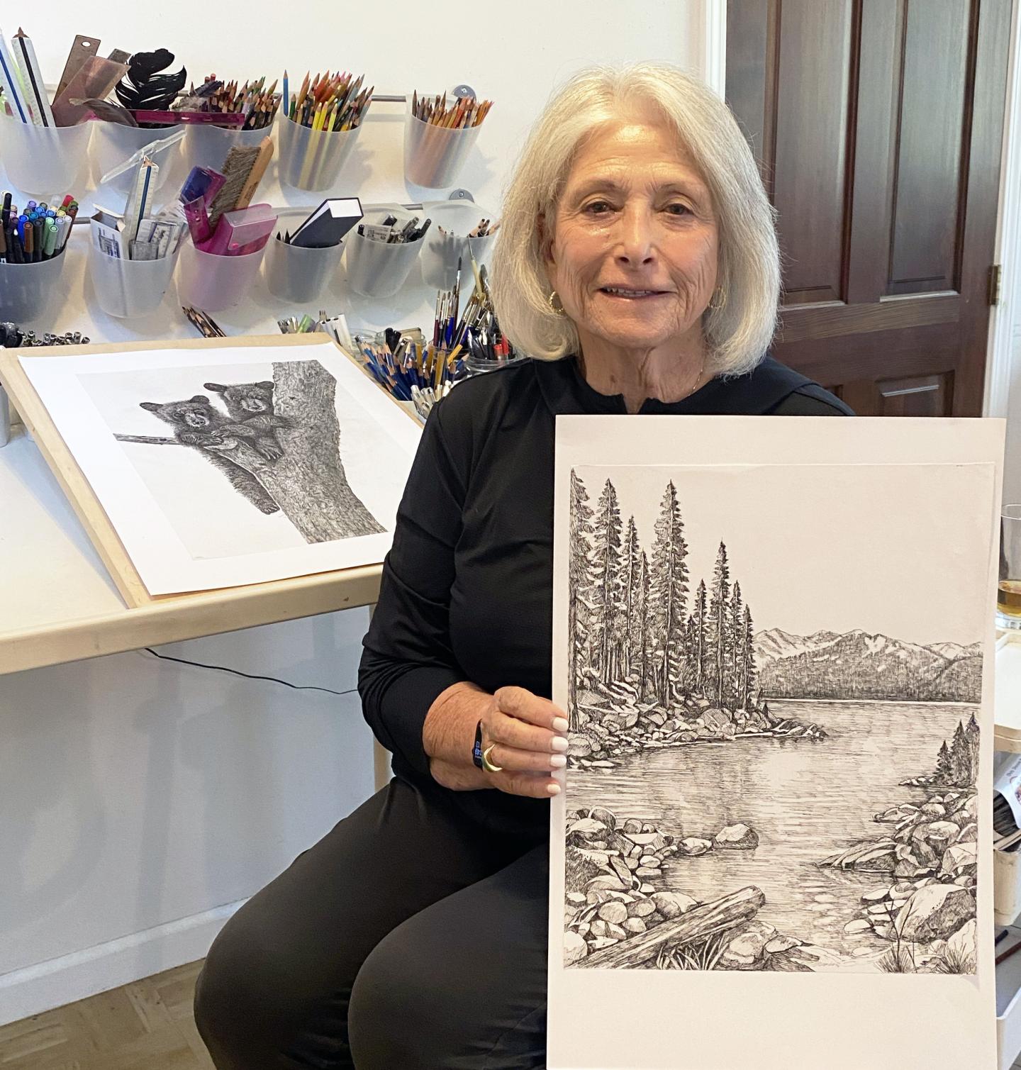 Woman holding a landscape drawing, surrounded by art supplies.