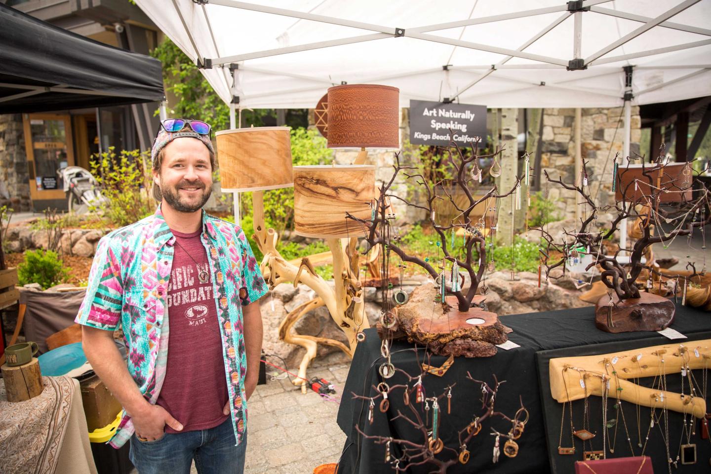 Man at an outdoor craft market stall with wooden art pieces.