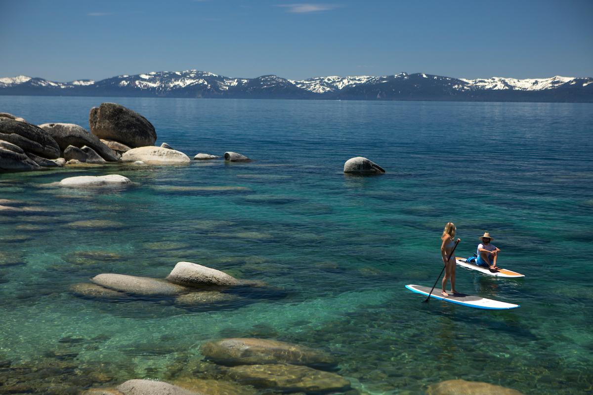 Paddleboarders on clear lake with distant snow-capped mountains.