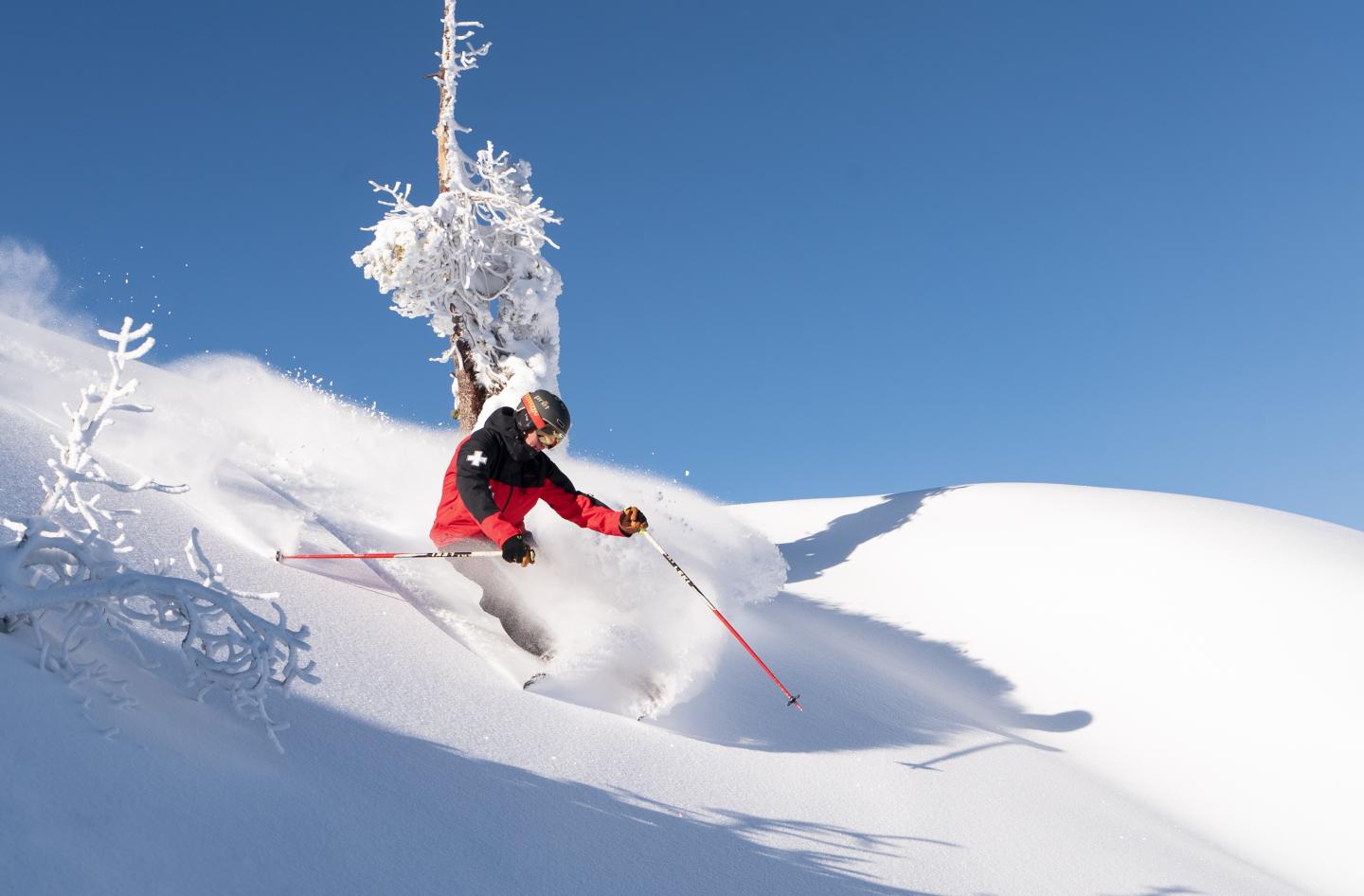 Skier in red jacket descending snowy slope under clear blue sky.