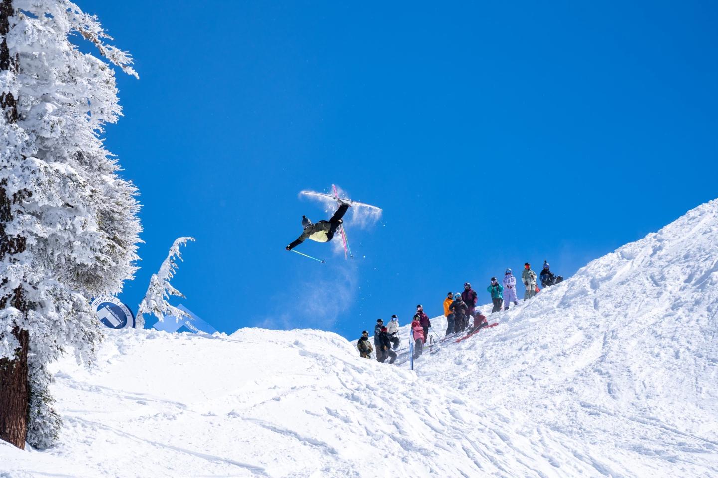 Skier jumps off snowy slope, group watches, clear blue sky. Silver Belt Classic at Sugar Bowl