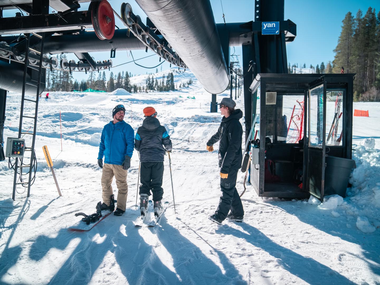 Skiers under a lift on a sunny snowy day, with pine trees in the background.