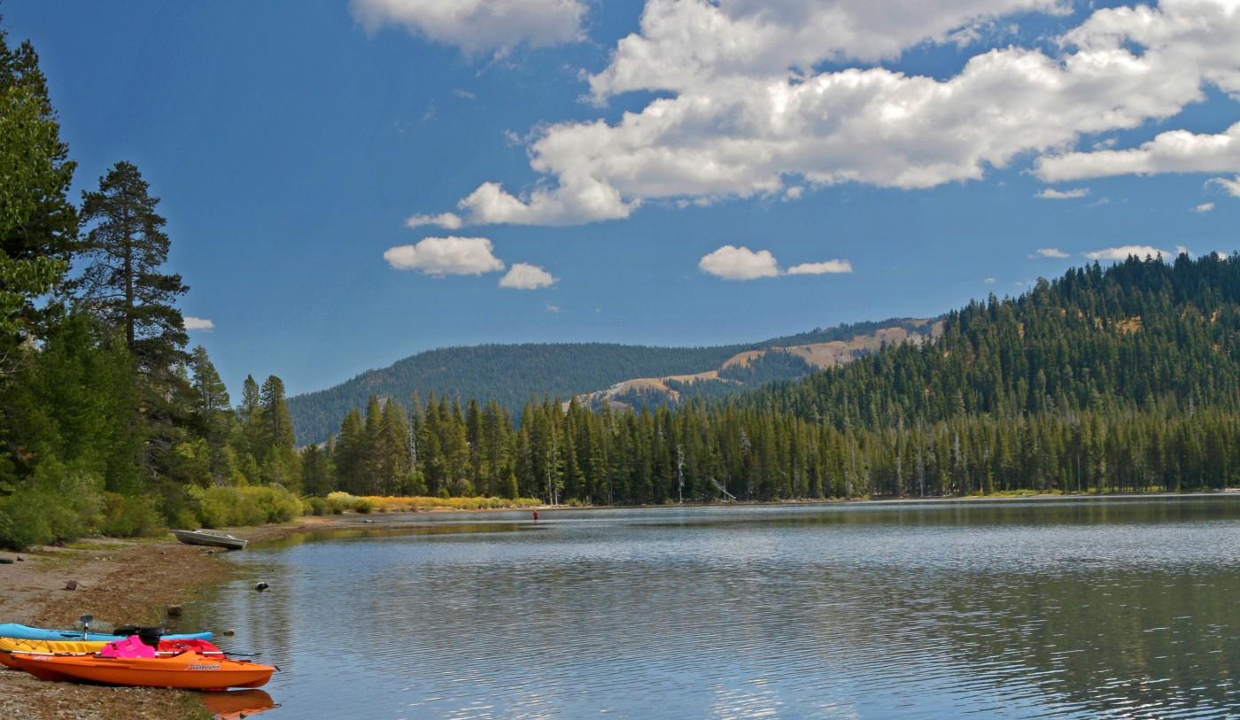 Mountain lake with kayaks, pine trees, and blue sky with clouds.