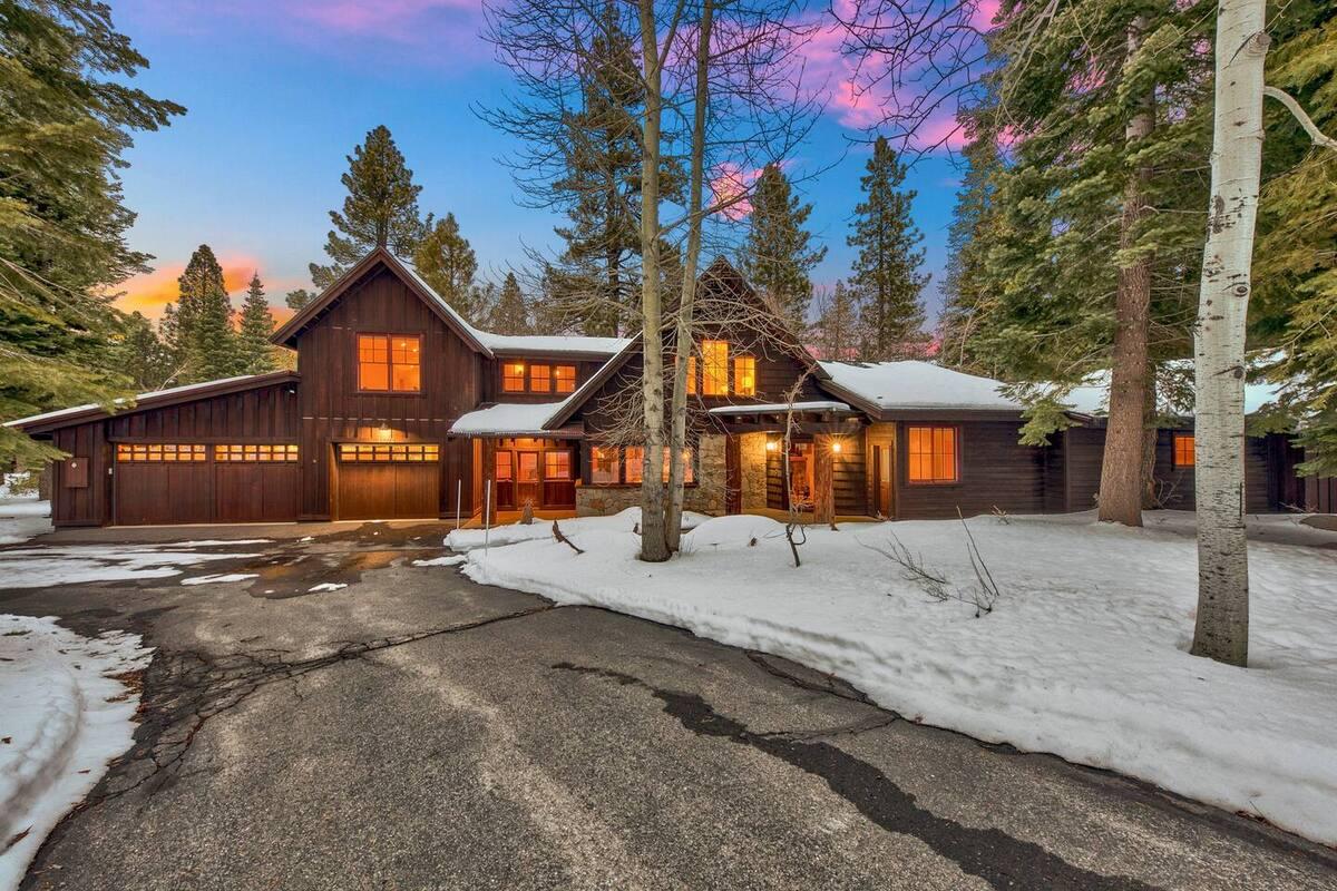 Snow-covered house surrounded by trees at sunset, warm lights inside.