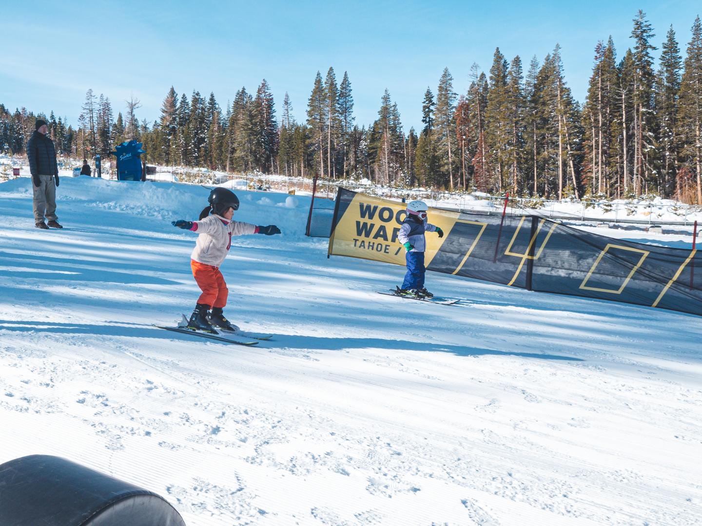 Children skiing on a snowy slope with trees and a banner in the background.