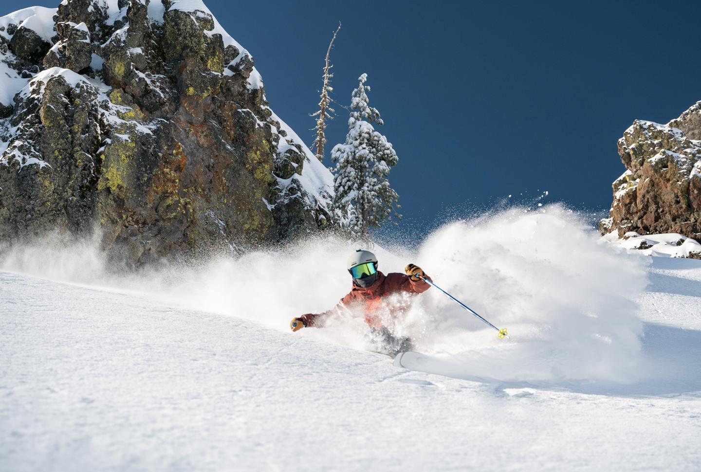 Skier in red jacket carving through powdery snow, rocky cliffside in background.