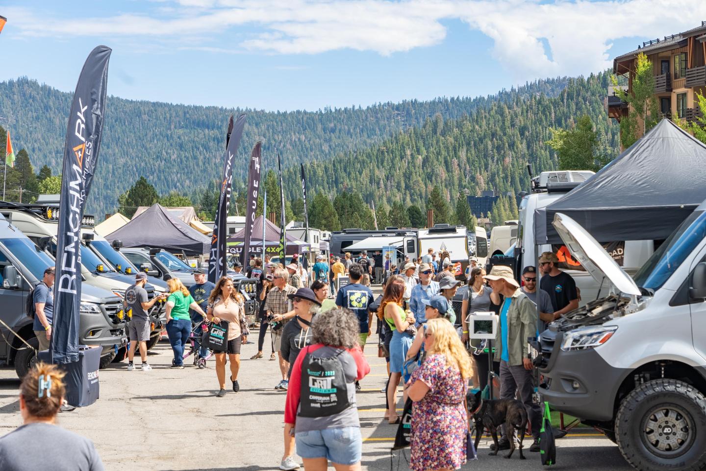 Outdoor festival with people, vans, and tents, surrounded by mountains.