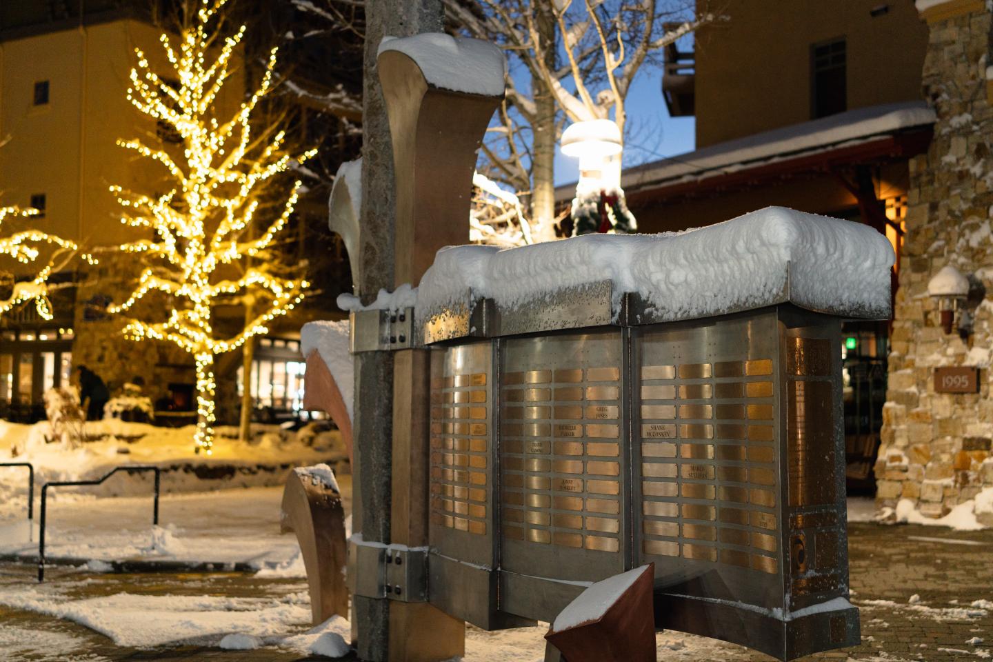 Snow-covered sculpture with lit trees in a winter evening setting.