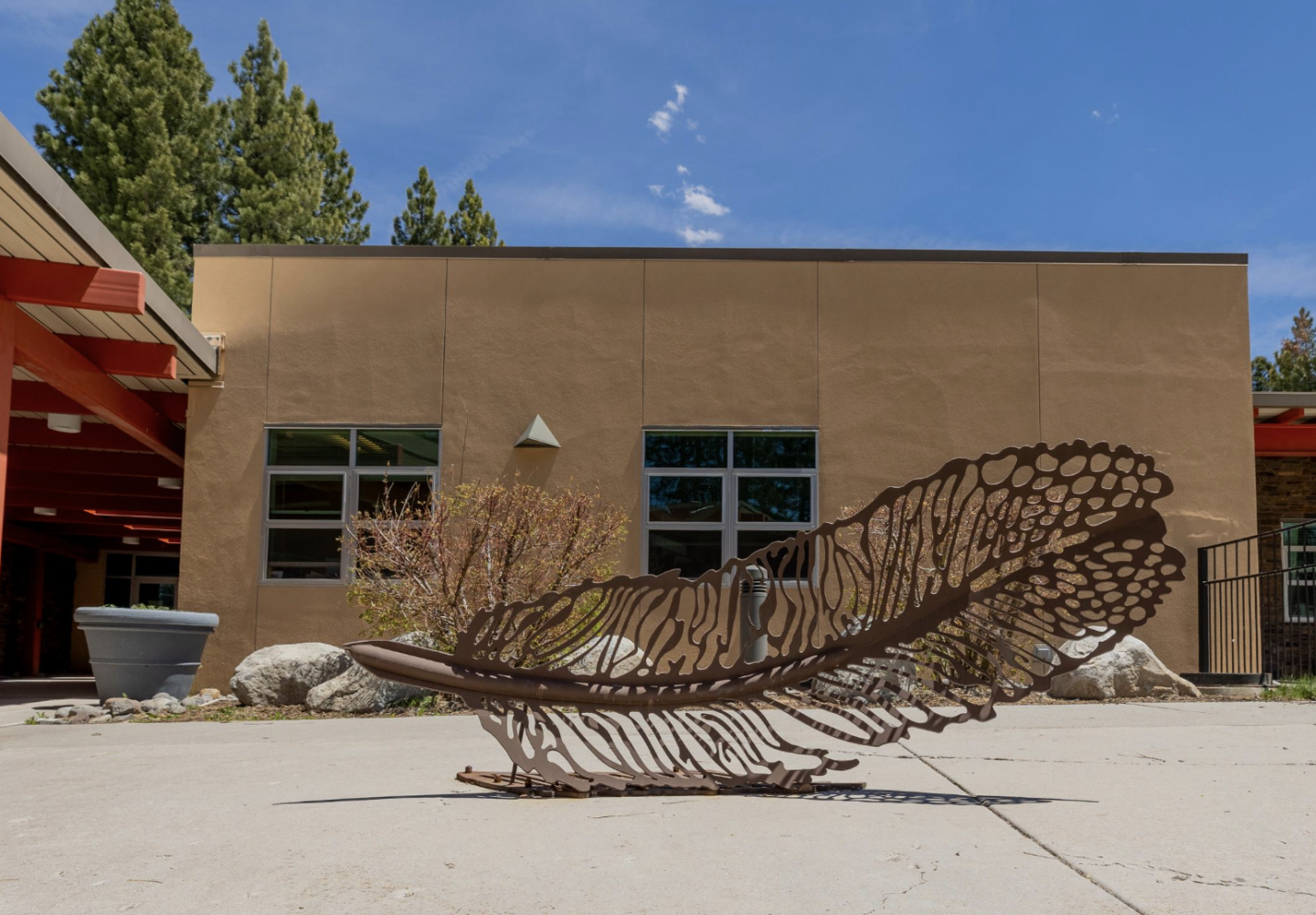 Large metal feather sculpture on concrete, modern building and trees in background.
