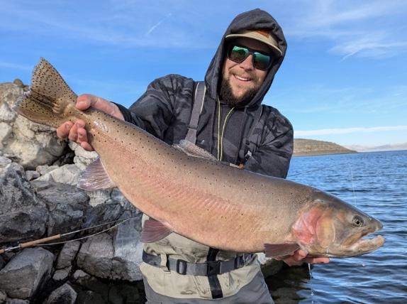 Man in sunglasses proudly holding a large fish by a lake.