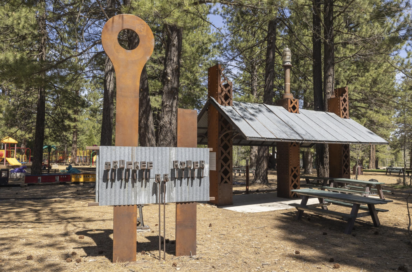 Outdoor pavilion with metallic sculptures and picnic tables among trees.